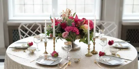A round wedding table set with a large pink and red floral centerpiece, tall white candles in gold holders, and gold-rimmed glassware.