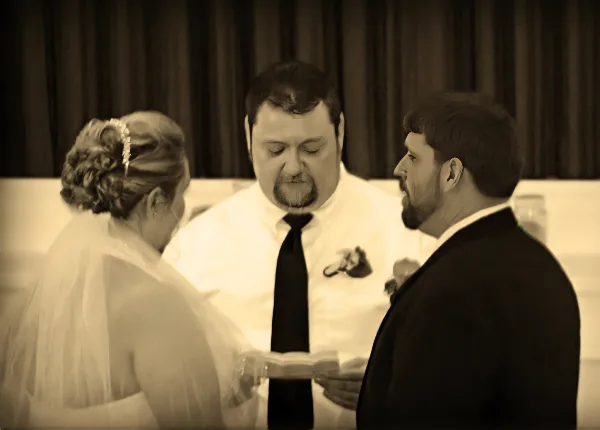 A wedding officiant in a white shirt and tie stands between a bride and groom during a ceremony.