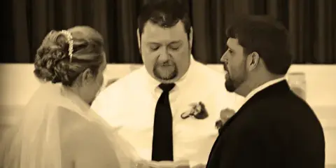 A wedding officiant in a white shirt and tie stands between a bride and groom during a ceremony.