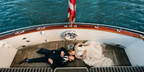 A bride and groom lying on the deck of a boat, viewed from above, with the ocean in the background.