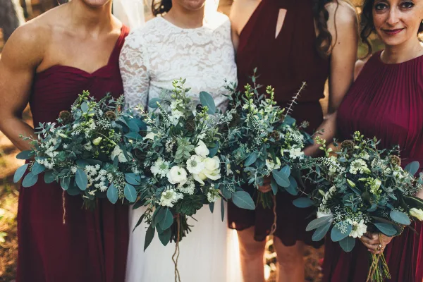 A bride and bridesmaids in maroon dresses holding lush, green and white floral bouquets.