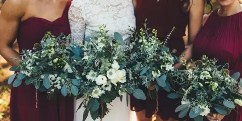 A bride and bridesmaids in maroon dresses holding lush, green and white floral bouquets.