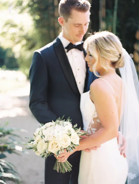 A bride and groom standing close together outdoors, with the bride holding a bouquet of white roses.
