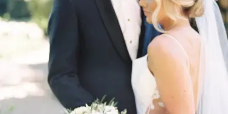 A bride and groom standing close together outdoors, with the bride holding a bouquet of white roses.