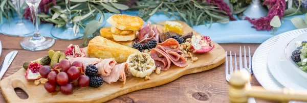 A wooden serving board filled with an assortment of cured meats, cheeses, grapes, blackberries, and crackers, set on a table with floral decorations.