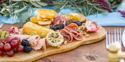 A wooden serving board filled with an assortment of cured meats, cheeses, grapes, blackberries, and crackers, set on a table with floral decorations.