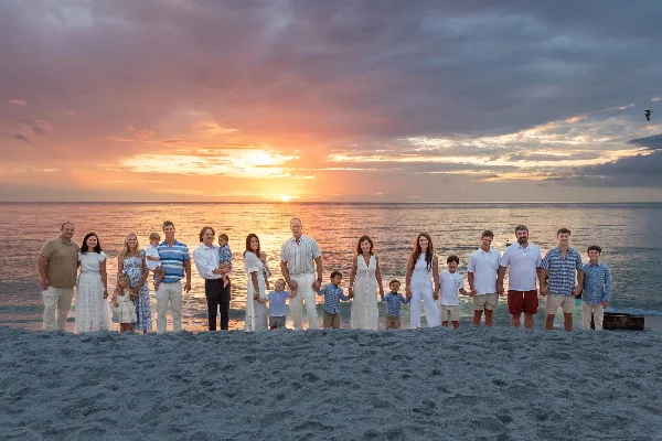 A large multi-generational family posing for a professional portrait on a sandy beach with a sunset over the ocean in the background.