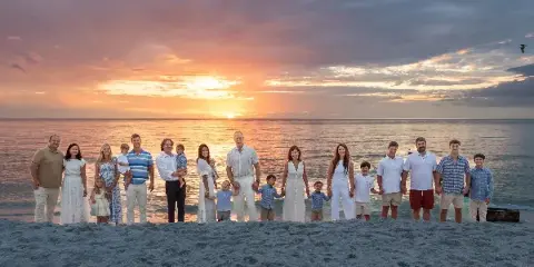 A large multi-generational family posing for a professional portrait on a sandy beach with a sunset over the ocean in the background.