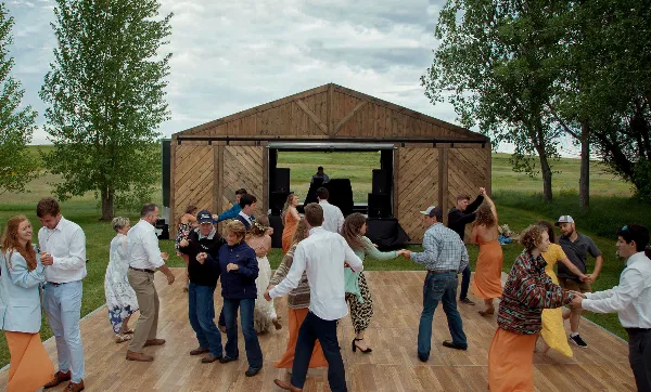 Guests dancing on a wooden floor in front of a mobile wooden barn venue in a grassy field.