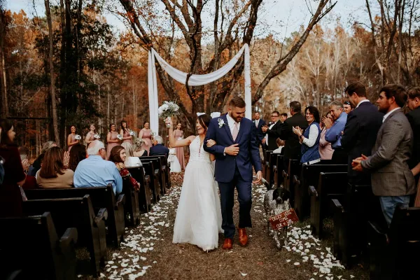 A bride and groom walking down the aisle during an outdoor wedding ceremony in a wooded area.