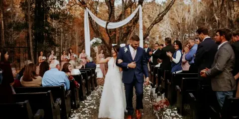 A bride and groom walking down the aisle during an outdoor wedding ceremony in a wooded area.