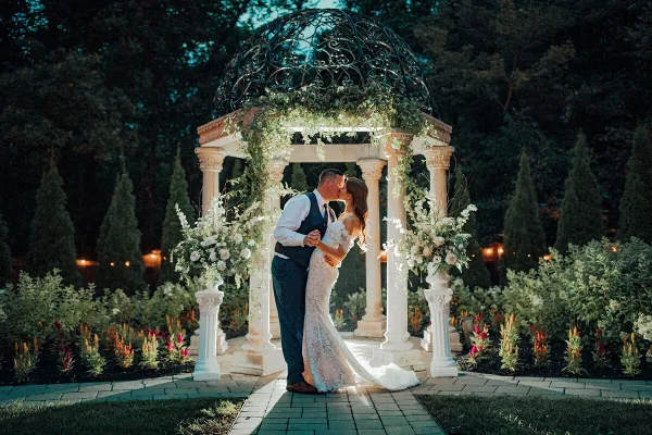 A bride and groom kissing under a decorative garden gazebo with floral arrangements.