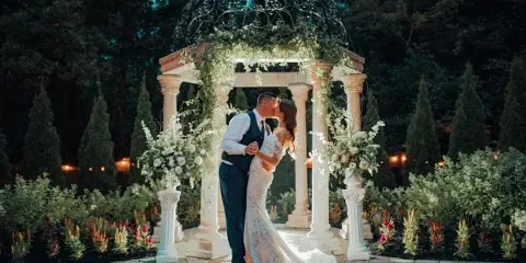 A bride and groom kissing under a decorative garden gazebo with floral arrangements.