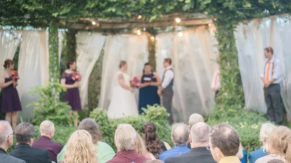 A wedding ceremony taking place outdoors under a leafy archway with a bride, groom, and officiant at the altar while guests watch.
