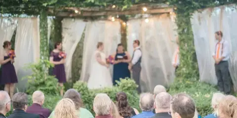 A wedding ceremony taking place outdoors under a leafy archway with a bride, groom, and officiant at the altar while guests watch.