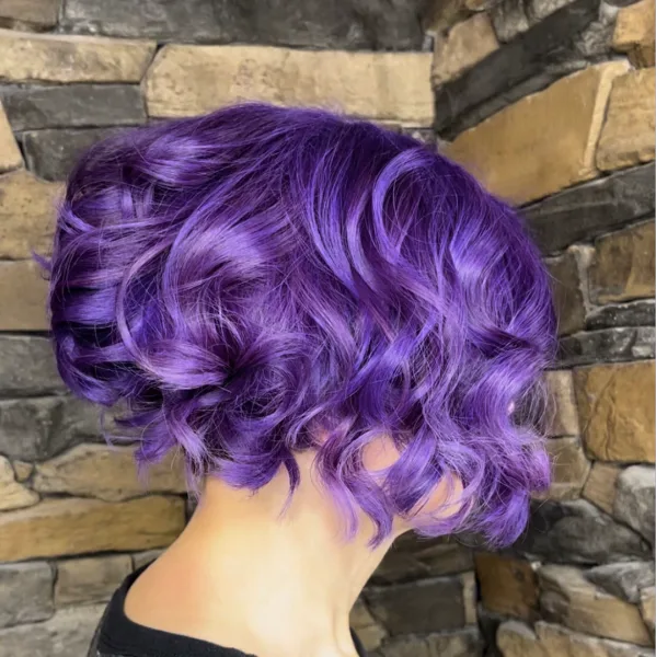 Back view of a person with a short, wavy purple hairstyle against a stone wall background.