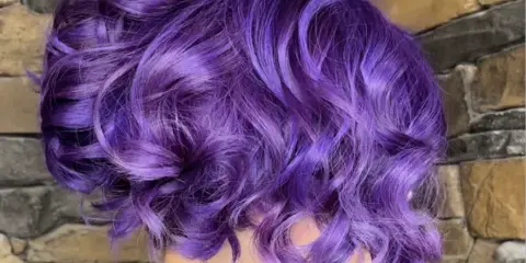 Back view of a person with a short, wavy purple hairstyle against a stone wall background.