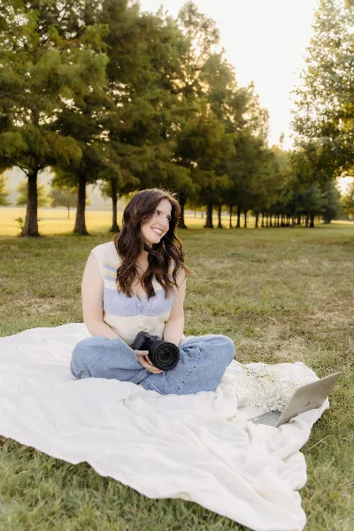 A woman sitting on a white blanket in a grassy field holding a black camera.
