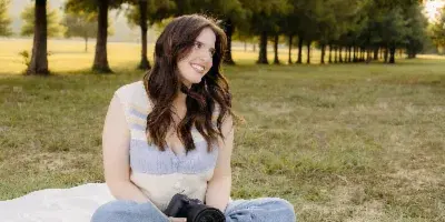 A woman sitting on a white blanket in a grassy field holding a black camera.