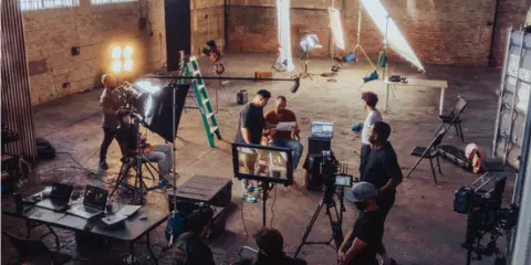 A film crew operating cameras and lighting equipment in a large studio space.