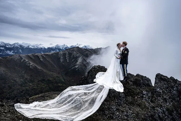 A bride and groom kissing on a rocky mountain peak with a long white veil flowing in the wind against a backdrop of misty mountains.