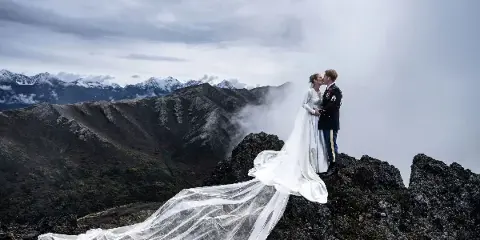 A bride and groom kissing on a rocky mountain peak with a long white veil flowing in the wind against a backdrop of misty mountains.