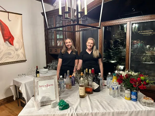 Two staff members in black uniforms standing behind a catering bar setup with liquor bottles, glasses, and an Izzy's Soirée sign.