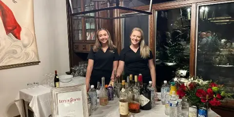 Two staff members in black uniforms standing behind a catering bar setup with liquor bottles, glasses, and an Izzy's Soirée sign.