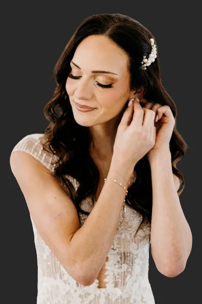 A bride with styled wavy hair and makeup, wearing a white lace dress and pearl hair accessory.