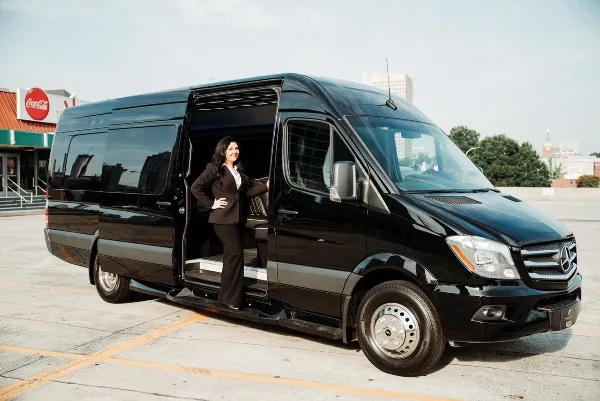 A black Mercedes-Benz Sprinter limousine van with a professional chauffeur standing in the open doorway.