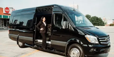 A black Mercedes-Benz Sprinter limousine van with a professional chauffeur standing in the open doorway.