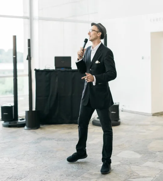 A man in a black suit and hat stands holding a microphone in a bright event venue with DJ equipment and speakers behind him.