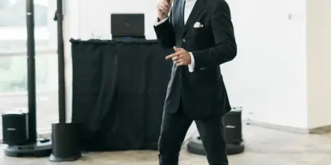 A man in a black suit and hat stands holding a microphone in a bright event venue with DJ equipment and speakers behind him.