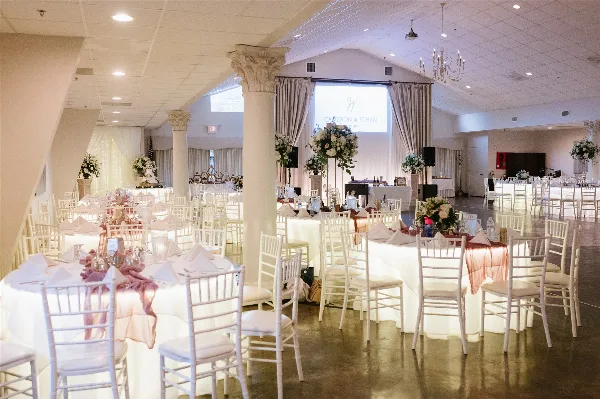 Interior view of a wedding venue featuring white columns, chandeliers, and round tables set with white linens and floral centerpieces.
