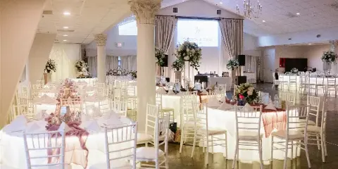 Interior view of a wedding venue featuring white columns, chandeliers, and round tables set with white linens and floral centerpieces.