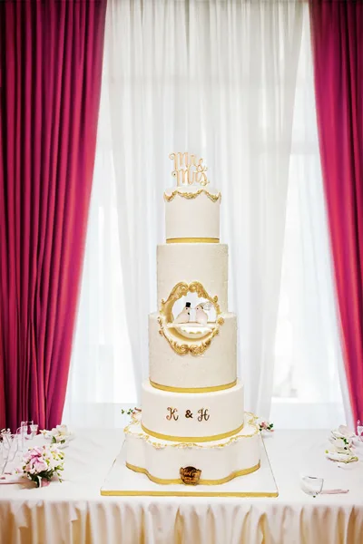 A four-tier white wedding cake with gold accents and a 'Mr & Mrs' topper displayed on a table.