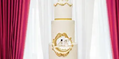 A four-tier white wedding cake with gold accents and a 'Mr & Mrs' topper displayed on a table.
