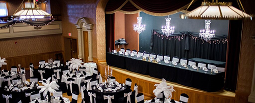 A formal event venue with a decorated stage, chandeliers, and banquet tables set with white floral centerpieces.