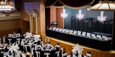 A formal event venue with a decorated stage, chandeliers, and banquet tables set with white floral centerpieces.