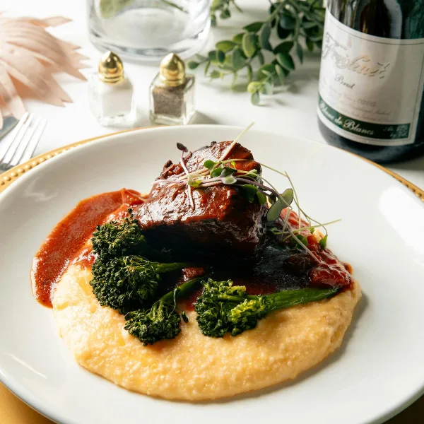 Plated gourmet dish with meat, broccoli, and sauce on a white plate with gold rim, set on a table with wine and greenery.