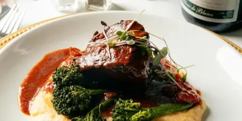 Plated gourmet dish with meat, broccoli, and sauce on a white plate with gold rim, set on a table with wine and greenery.