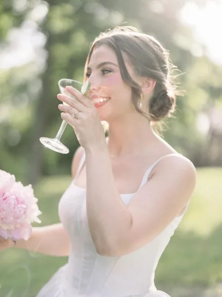 A bride in a white dress with styled hair and makeup holding a glass and a bouquet.