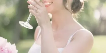 A bride in a white dress with styled hair and makeup holding a glass and a bouquet.
