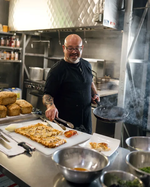 Chef in a professional kitchen plating a prepared dish with a pan and tongs, with loaves of bread and bowls of ingredients nearby.