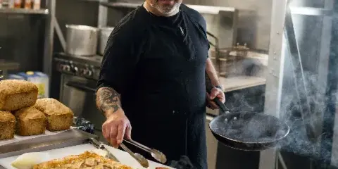 Chef in a professional kitchen plating a prepared dish with a pan and tongs, with loaves of bread and bowls of ingredients nearby.