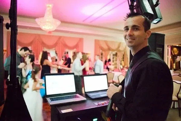 A professional DJ at a wedding reception with a laptop setup, while a bride and groom dance in the background.