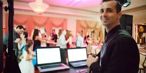 A professional DJ at a wedding reception with a laptop setup, while a bride and groom dance in the background.
