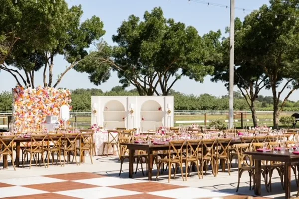 Outdoor event setup with long wooden tables, cross-back chairs, a white lounge structure, and floral arrangements.