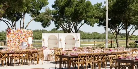 Outdoor event setup with long wooden tables, cross-back chairs, a white lounge structure, and floral arrangements.
