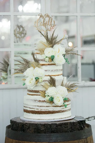 A three-tiered semi-naked wedding cake decorated with white roses, dried grasses, and a gold topper, displayed on a wooden barrel stand.